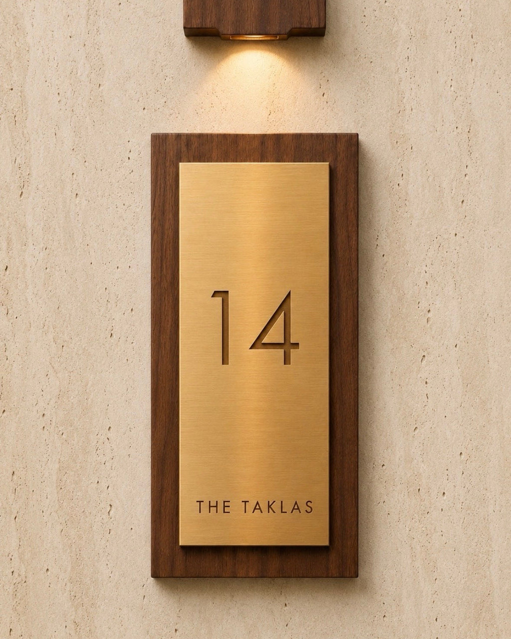 A custom house number plaque featuring a brushed brass plate', mounted in a dark wood frame on a textured beige wall under a matching wall sconce.
