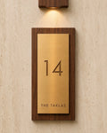 A custom house number plaque featuring a brushed brass plate', mounted in a dark wood frame on a textured beige wall under a matching wall sconce.