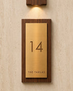 A custom house number plaque featuring a brushed brass plate', mounted in a dark wood frame on a textured beige wall under a matching wall sconce.
