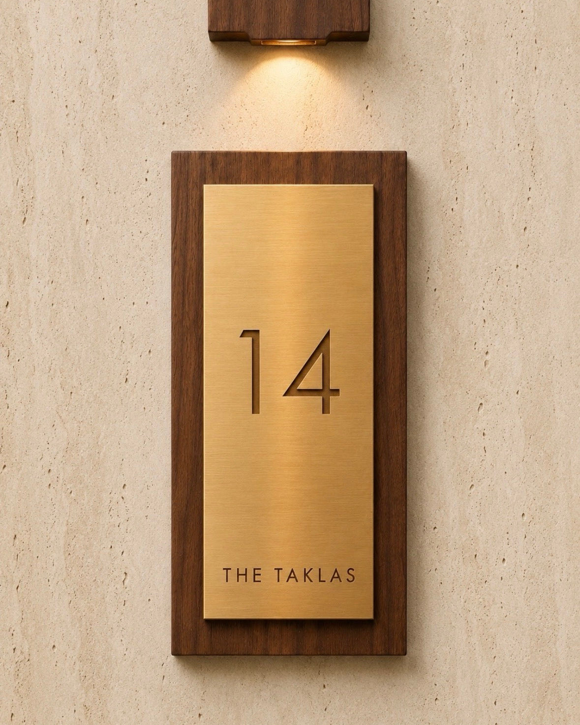 A custom house number plaque featuring a brushed brass plate', mounted in a dark wood frame on a textured beige wall under a matching wall sconce.
